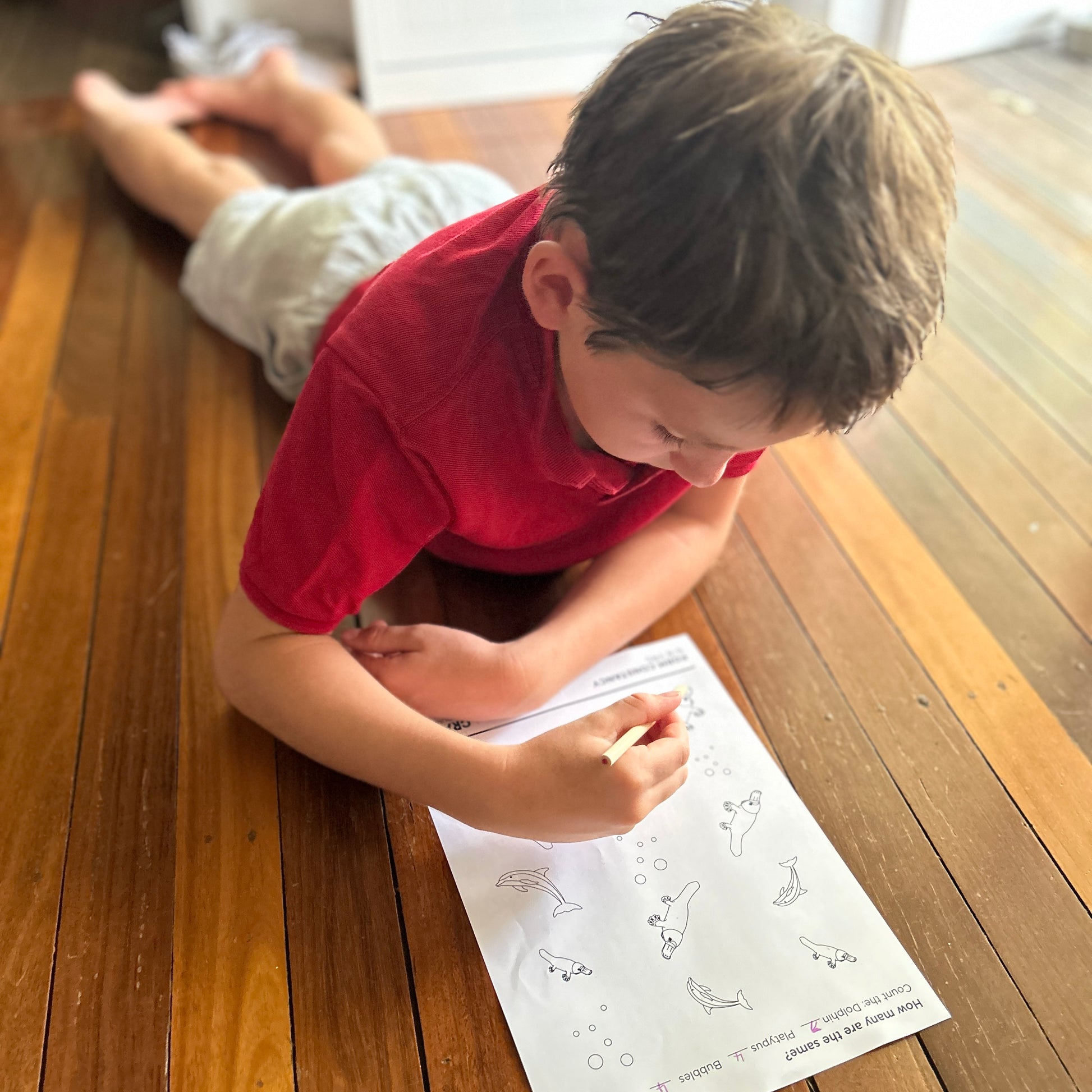 Young boy in red shirt lying on floor with pencil and worksheet